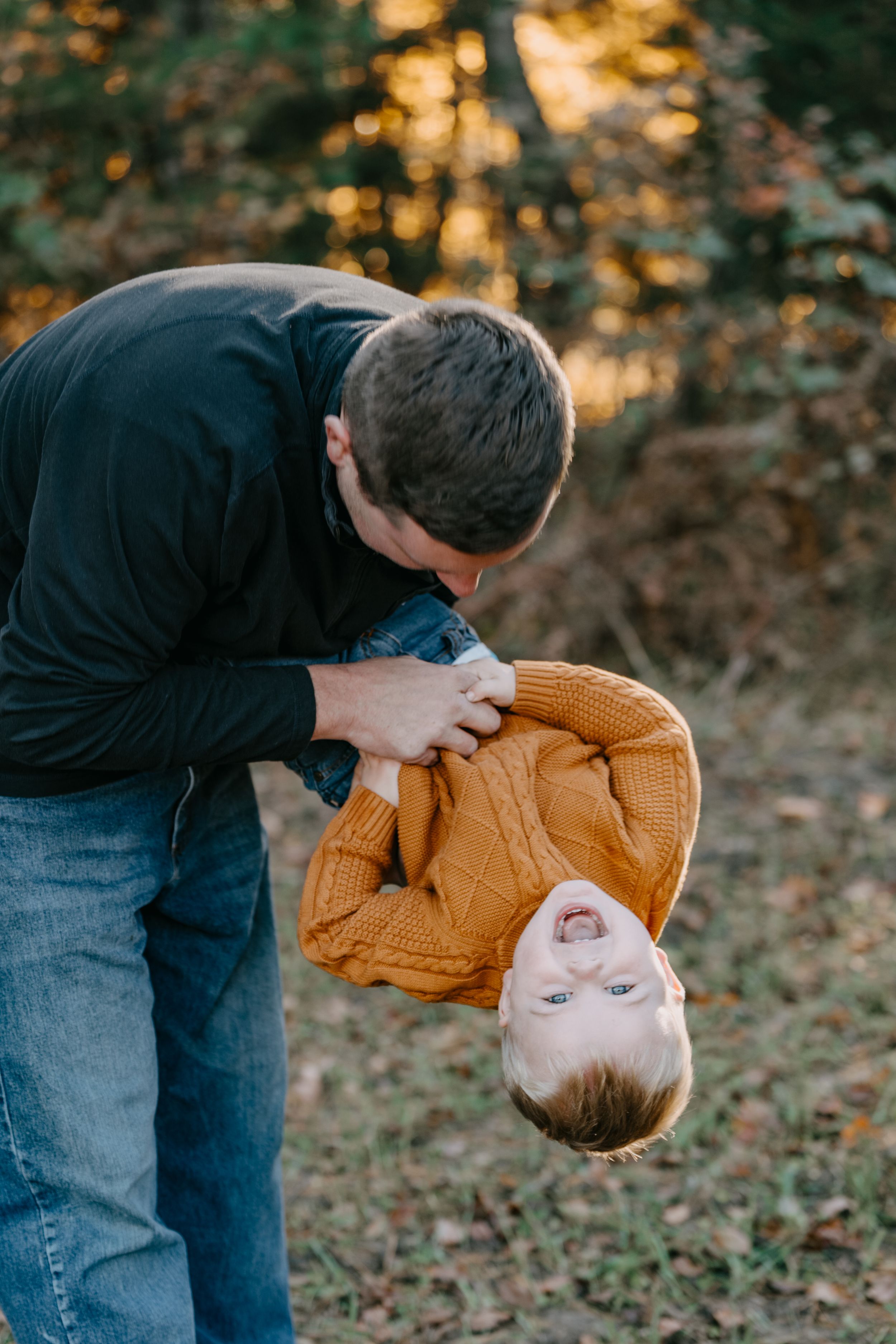 Person in black shirt and jeans bends over to pick up or set down a brown pumpkin on autumn ground covered in leaves.