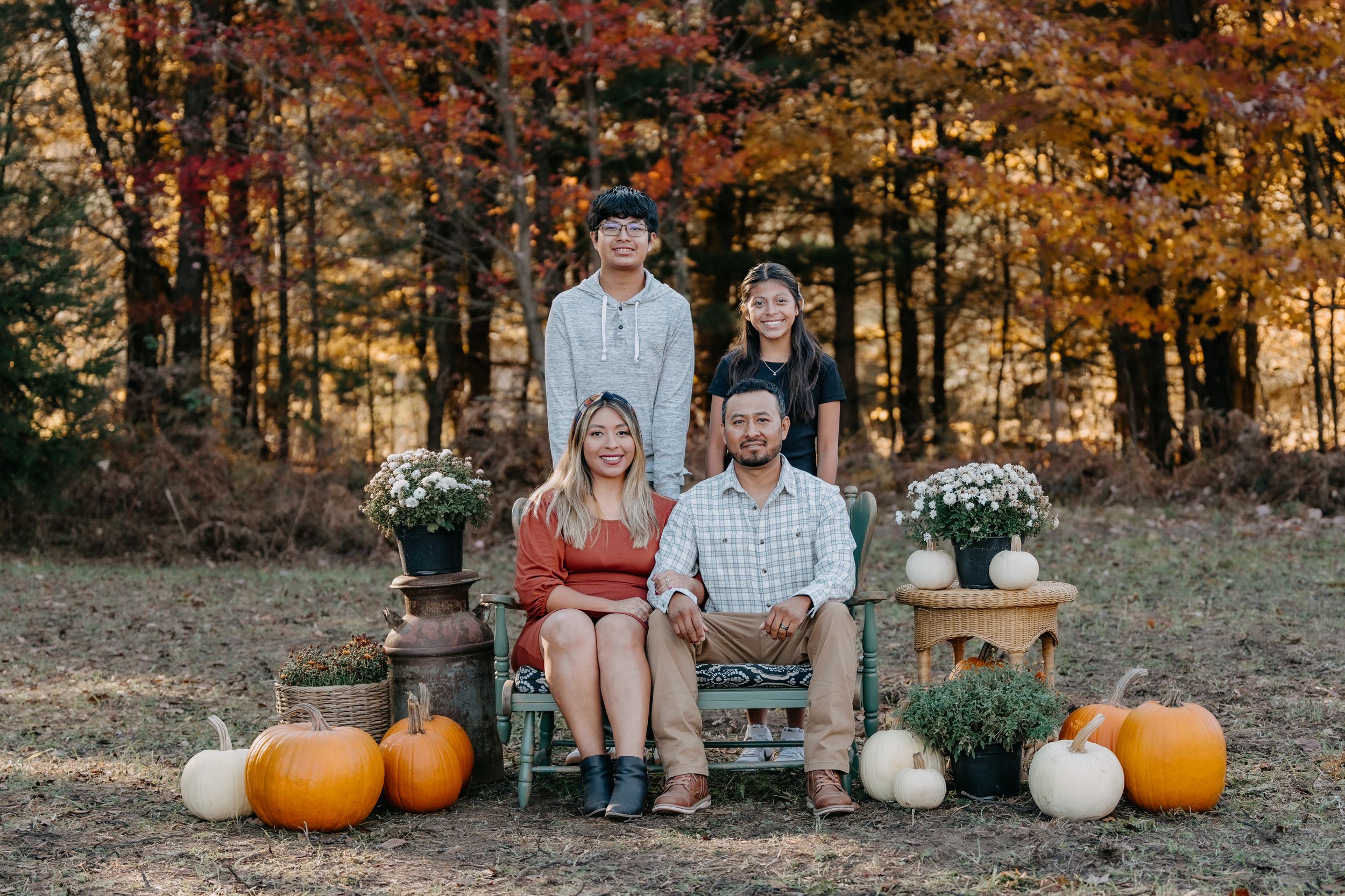 Seasonal autumn outdoor portrait of a family sitting on rustic bench with orange pumpkins and white mum flowers in woods.