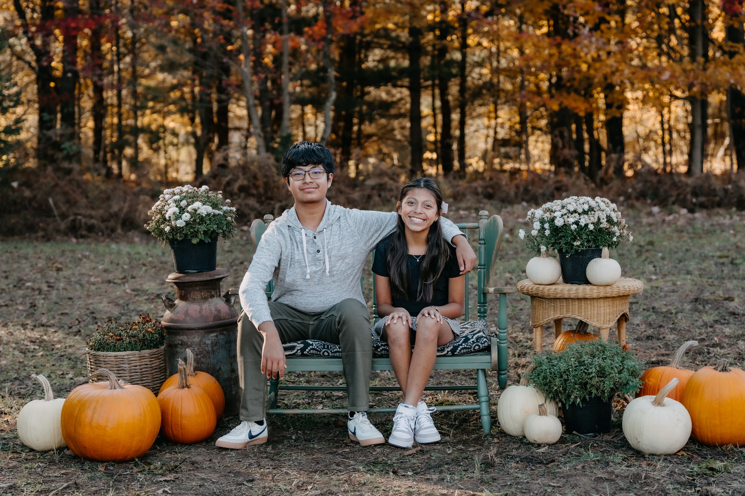 Two people sit on a rustic outdoor bench surrounded by autumn pumpkins and white flowers in a fall forest setting.