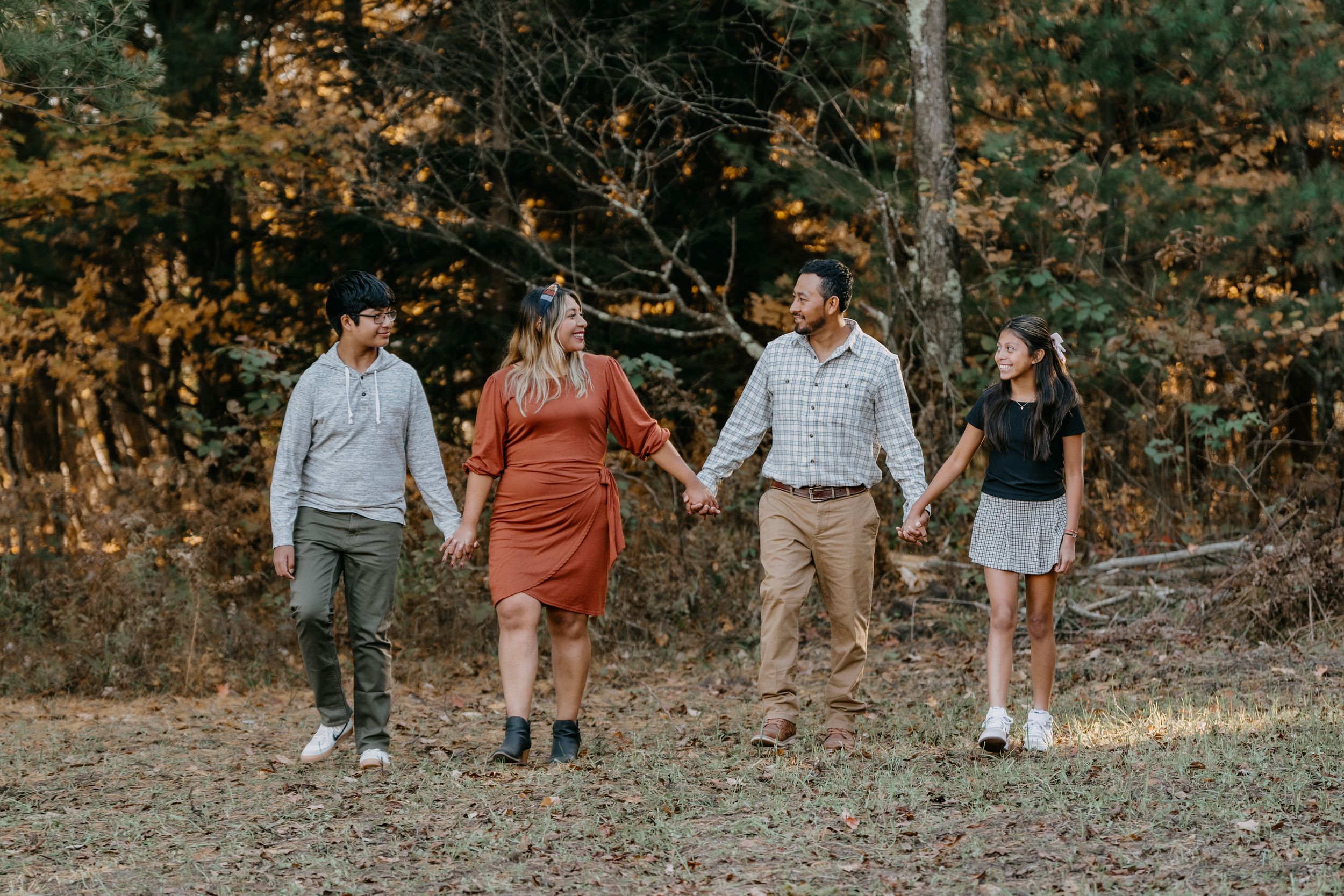 A group of people walk and laugh together along a wooded path during golden hour in autumn colors.