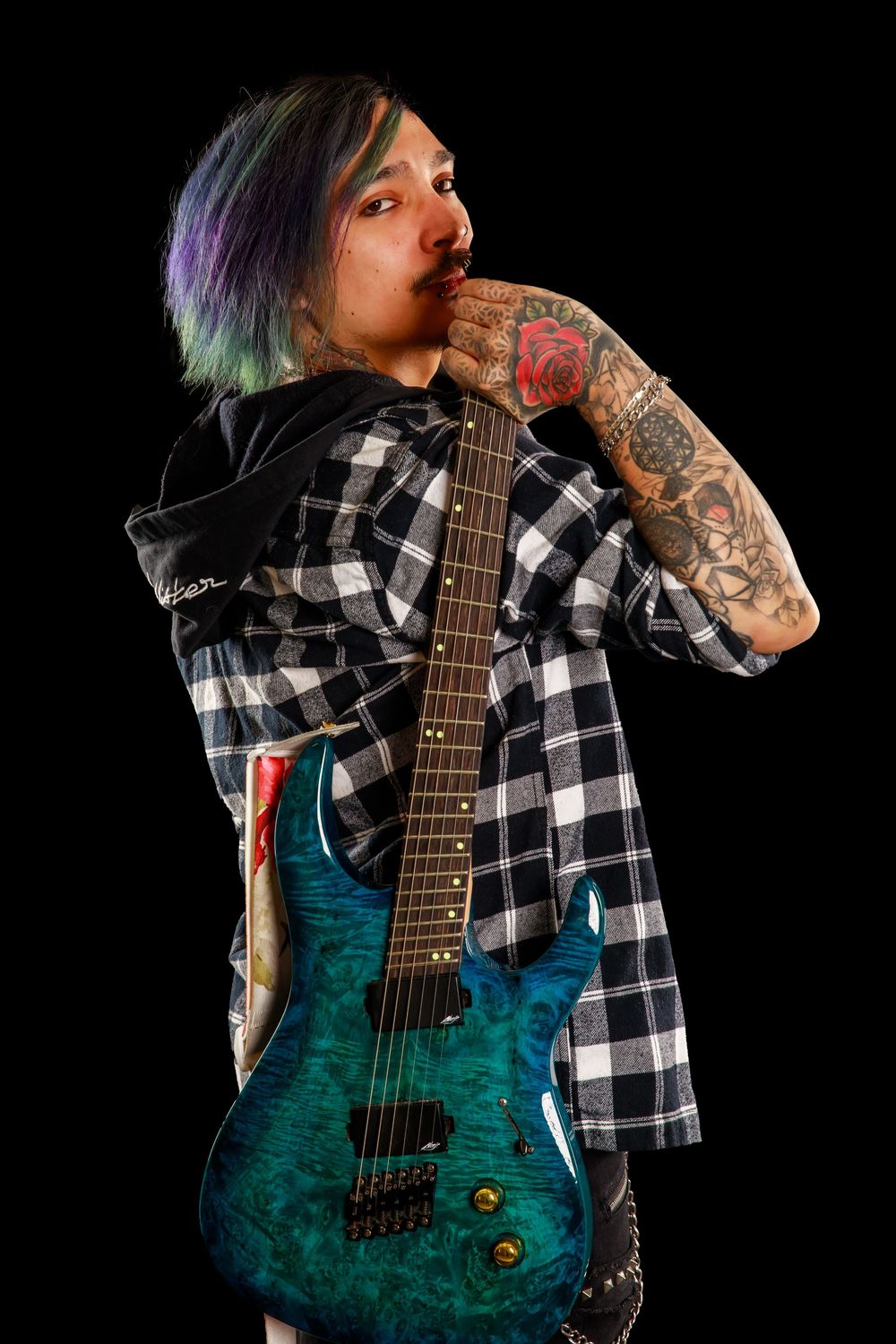 Musician in plaid flannel shirt plays teal electric guitar against dark background.