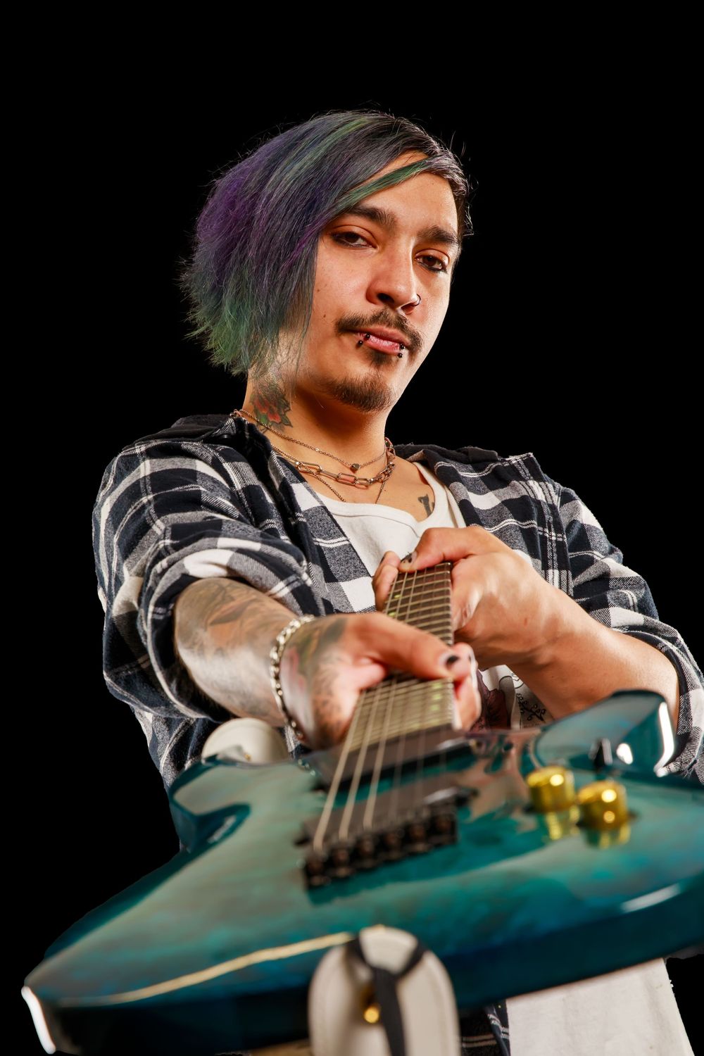Close-up shot of guitarist with colorful hair playing teal electric guitar while wearing plaid shirt.