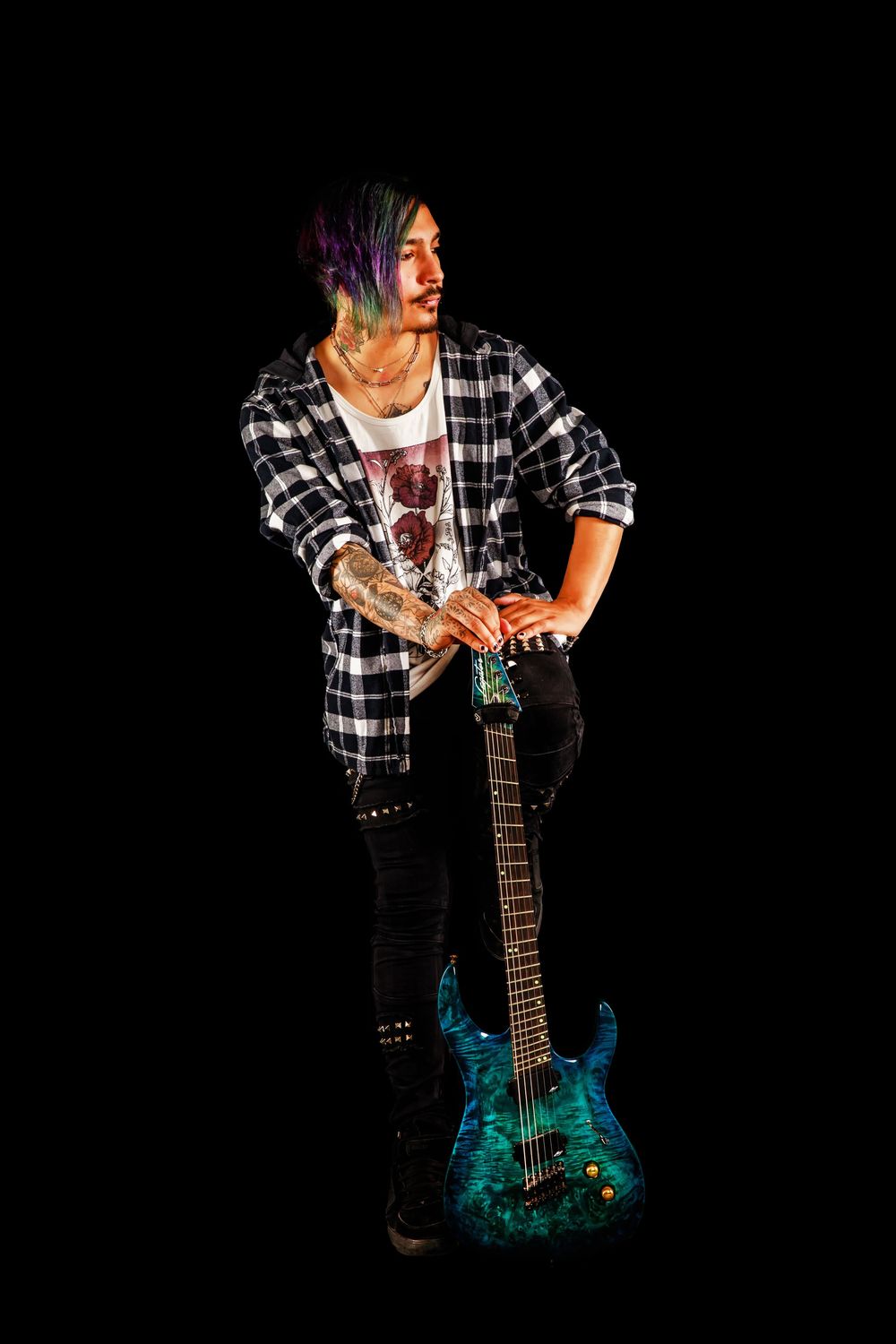 Musician in plaid flannel and dark jeans poses with a teal electric guitar against black background.