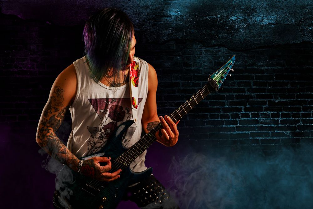 A tattooed musician plays electric guitar in dramatic lighting against a dark brick wall backdrop.
