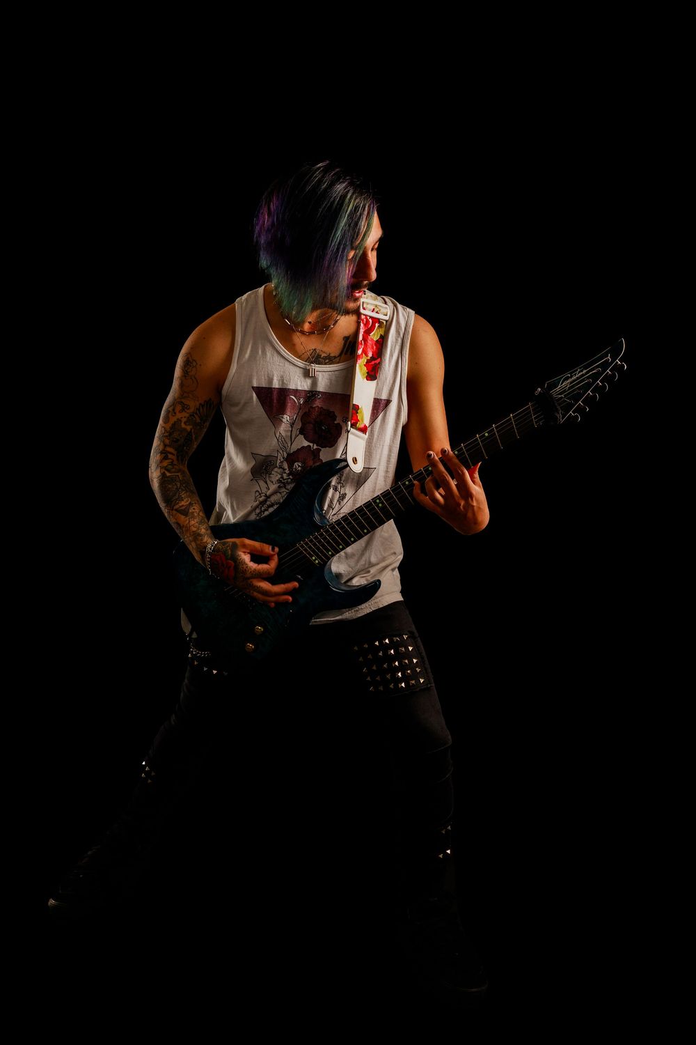 Tattooed guitarist in white tank top performs on dark stage with dramatic lighting.