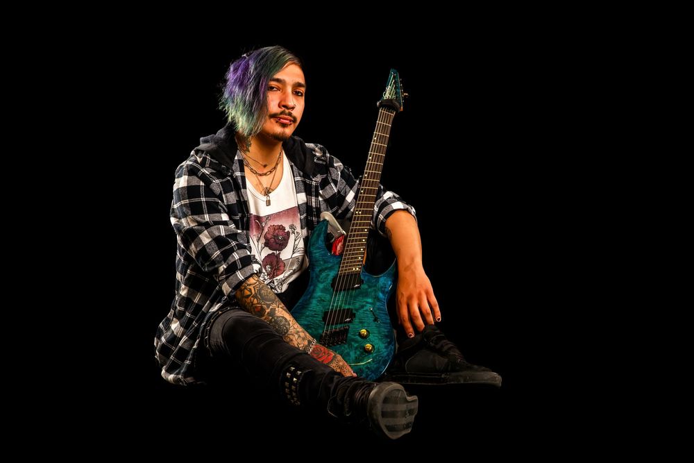 A musician in a plaid flannel shirt sits with their electric guitar against a black background during a photo shoot.