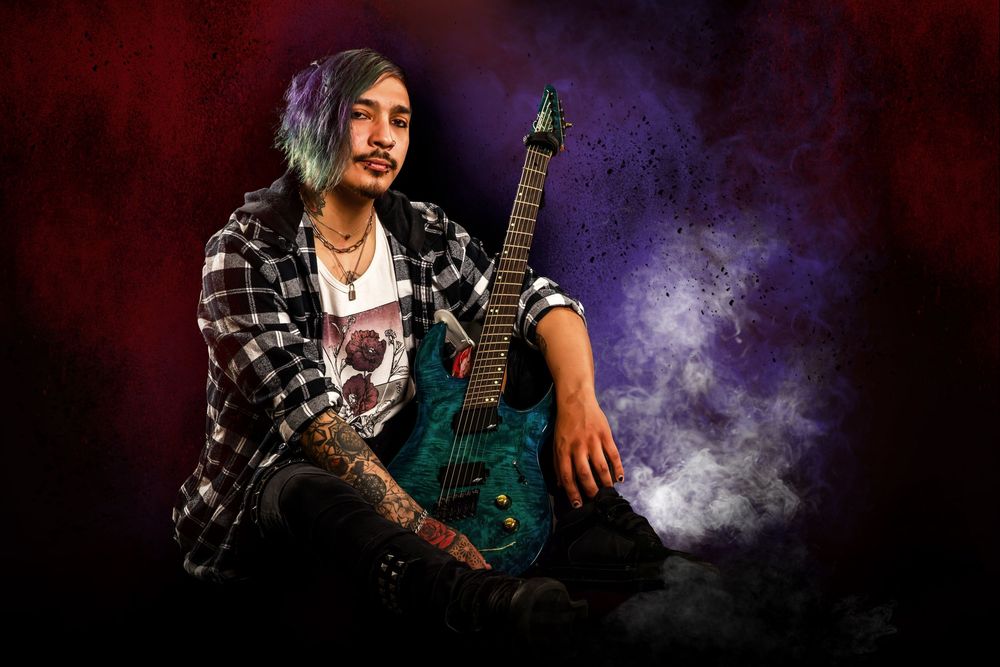 Alternative style musician poses with turquoise guitar against smoky purple and red backdrop.