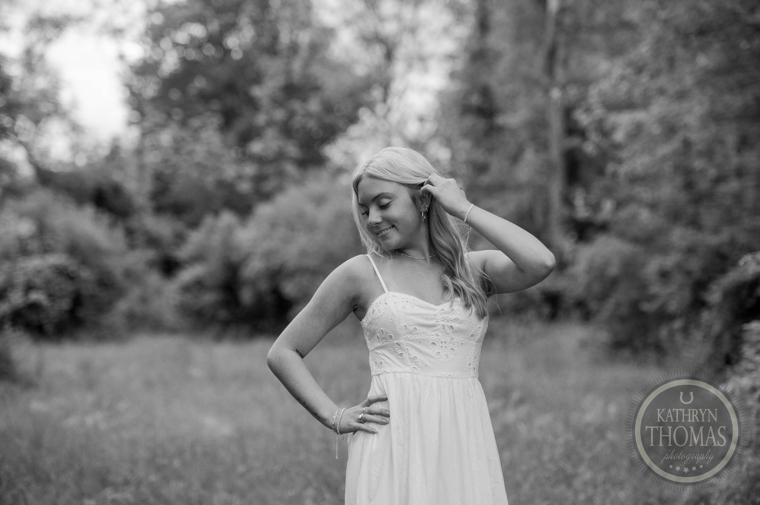 Woman wearing a white summer dress smiles while standing in a green meadow on a sunny afternoon.