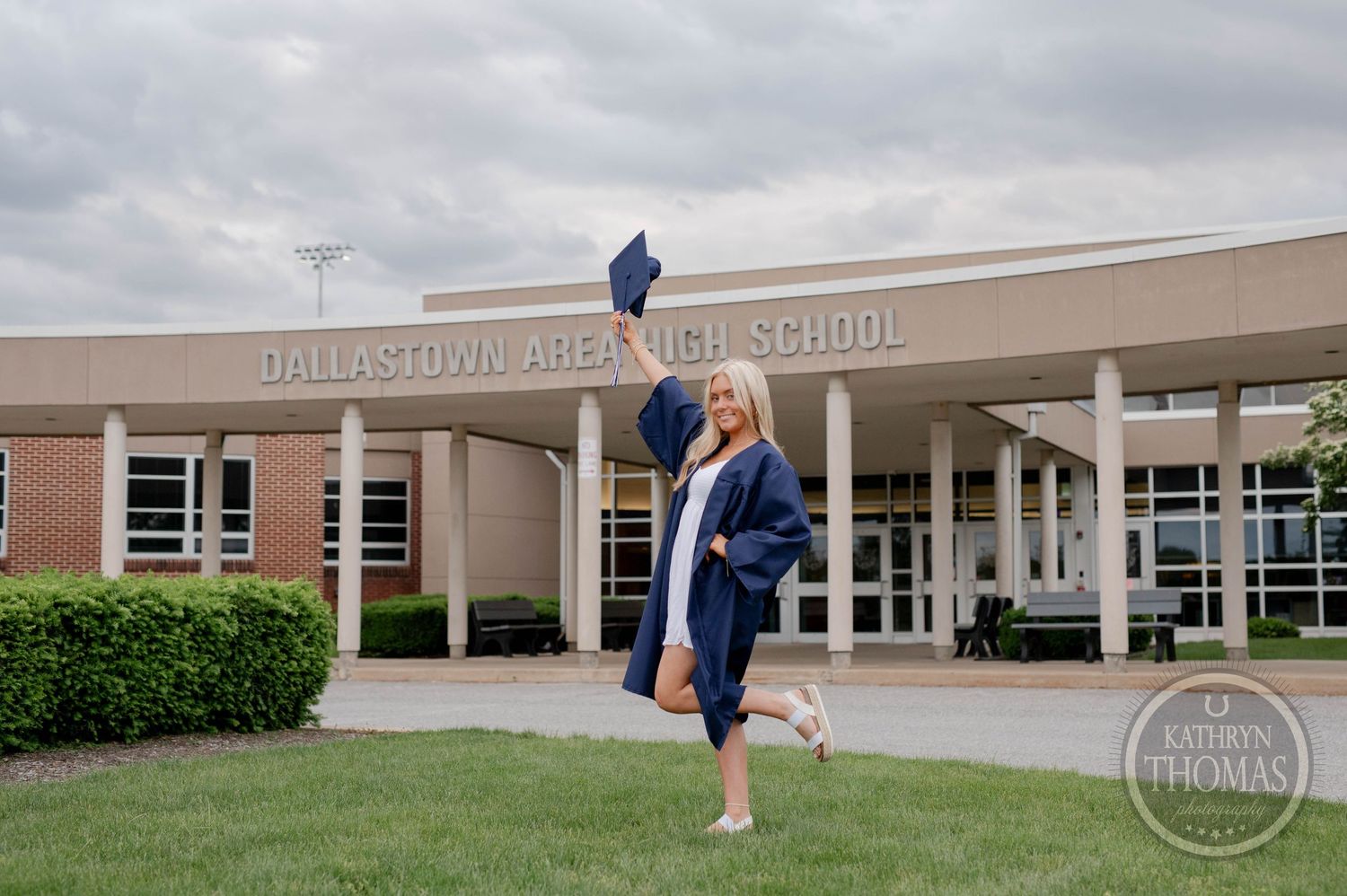 Students in dark blue graduation gowns gather outside a high school building for graduation photos with balloons.