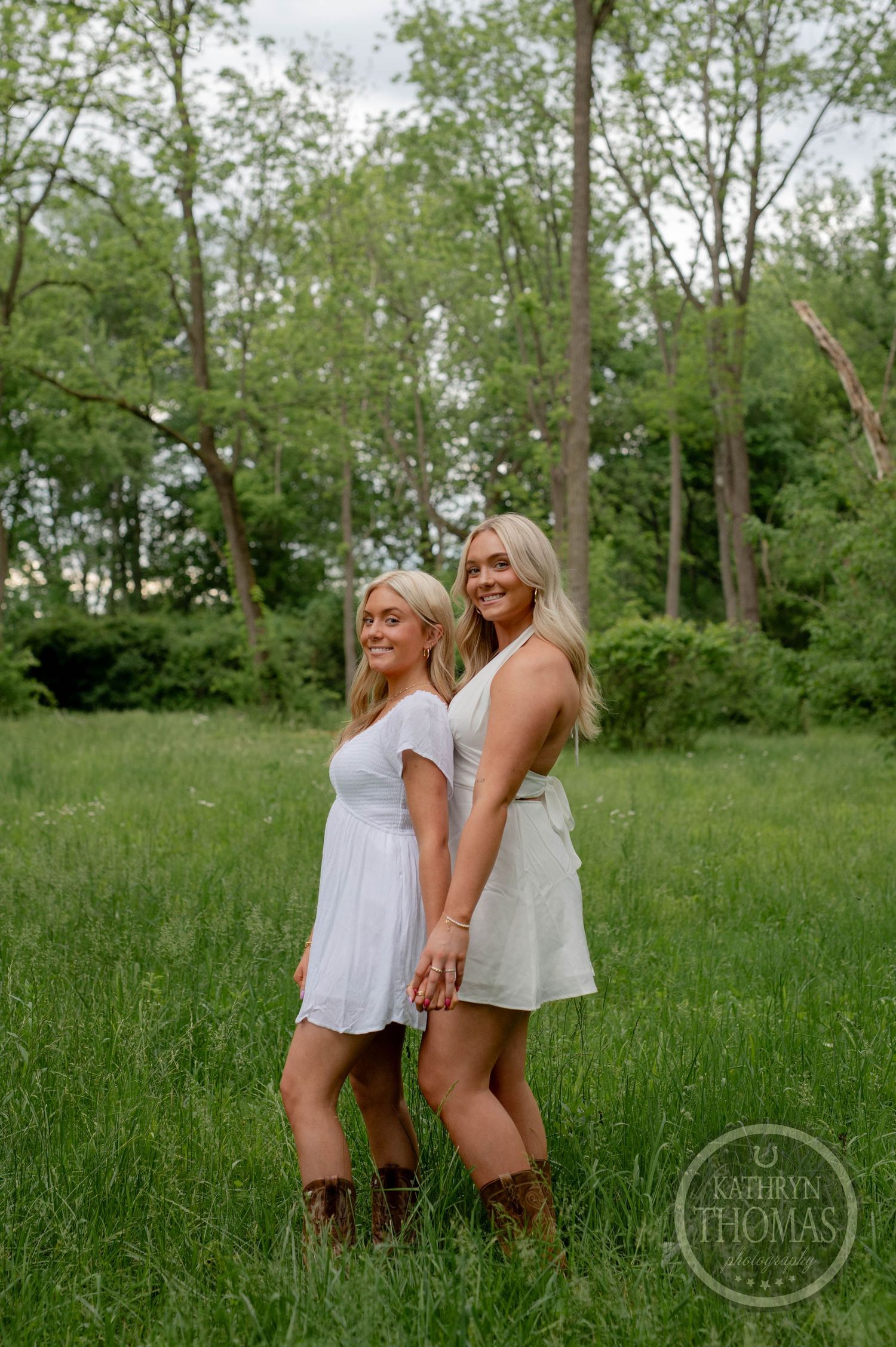 Two people in white dresses stand together in a grassy field with brown boots, surrounded by green trees.