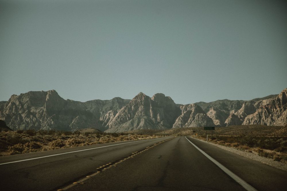 A winding asphalt road stretches through rugged desert terrain towards distant rocky mountains under a hazy sky.