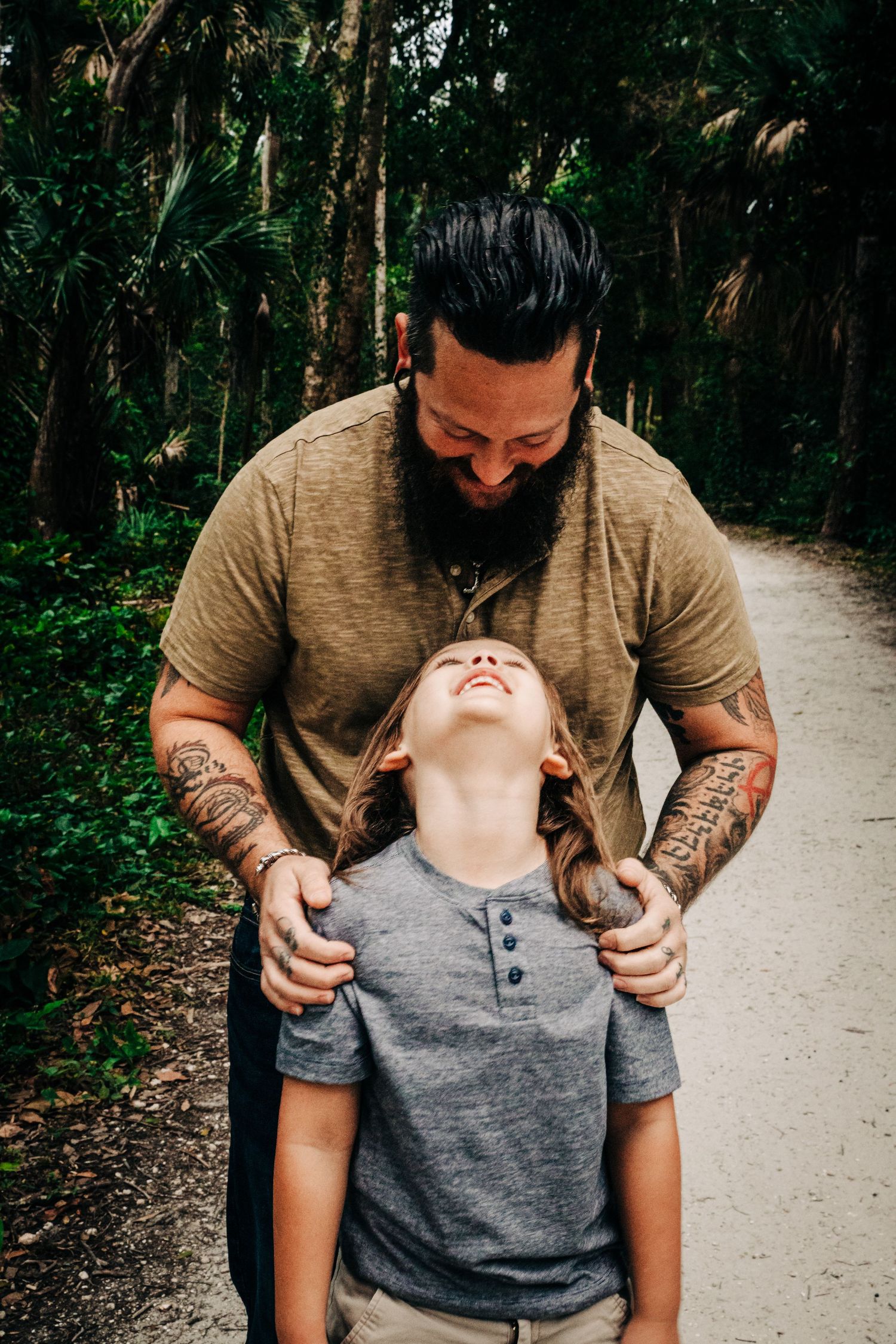 On a wooded trail, a child and adult wearing tan and gray shirts share a joyful, candid moment together at riverbend park in Jupiter fl.