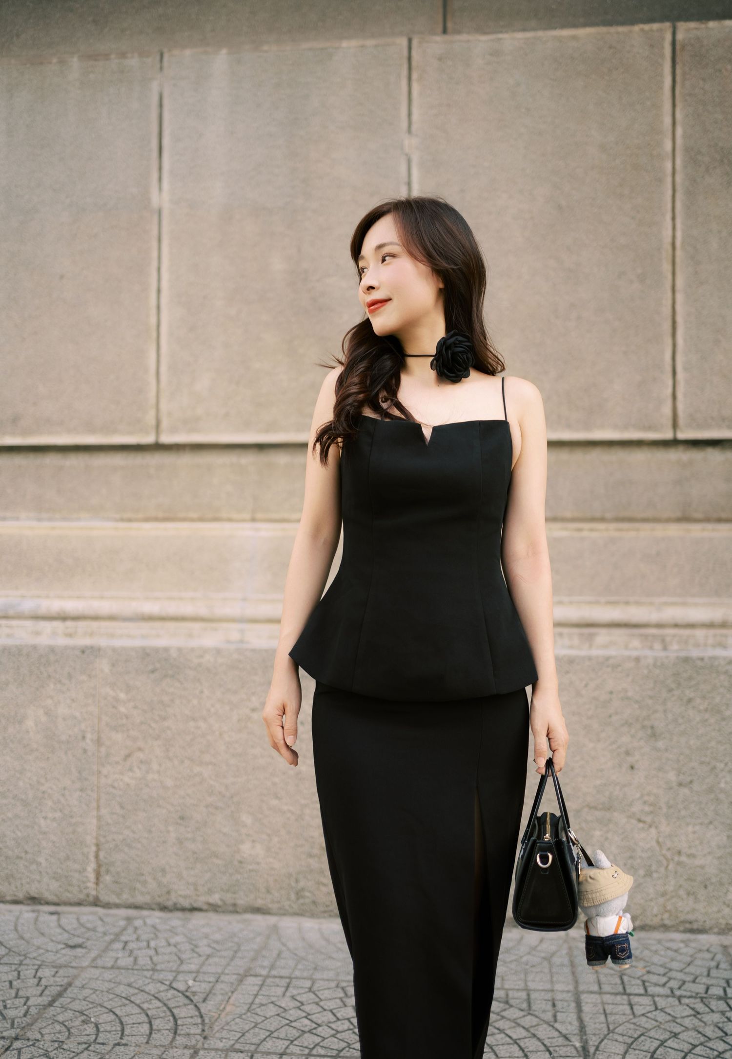 A person wearing a long black sleeveless dress stands against a concrete wall while holding a small handbag.