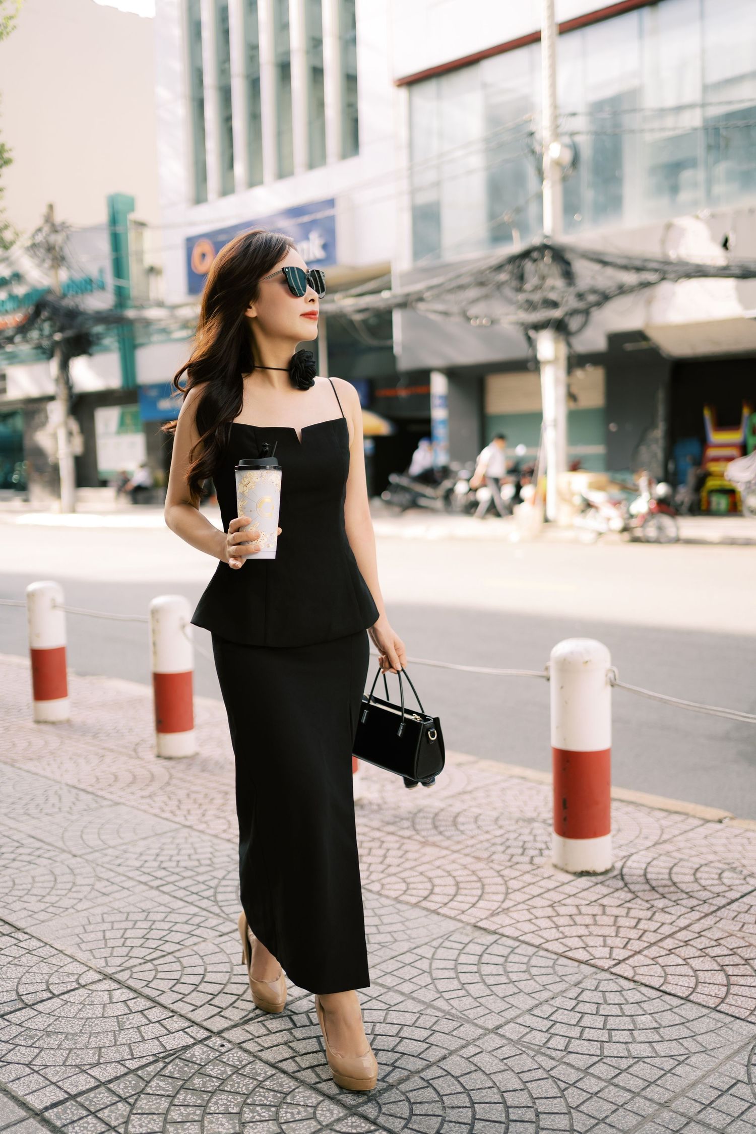 Stylish woman in black dress holding coffee walking on urban sidewalk with red and white bollards.