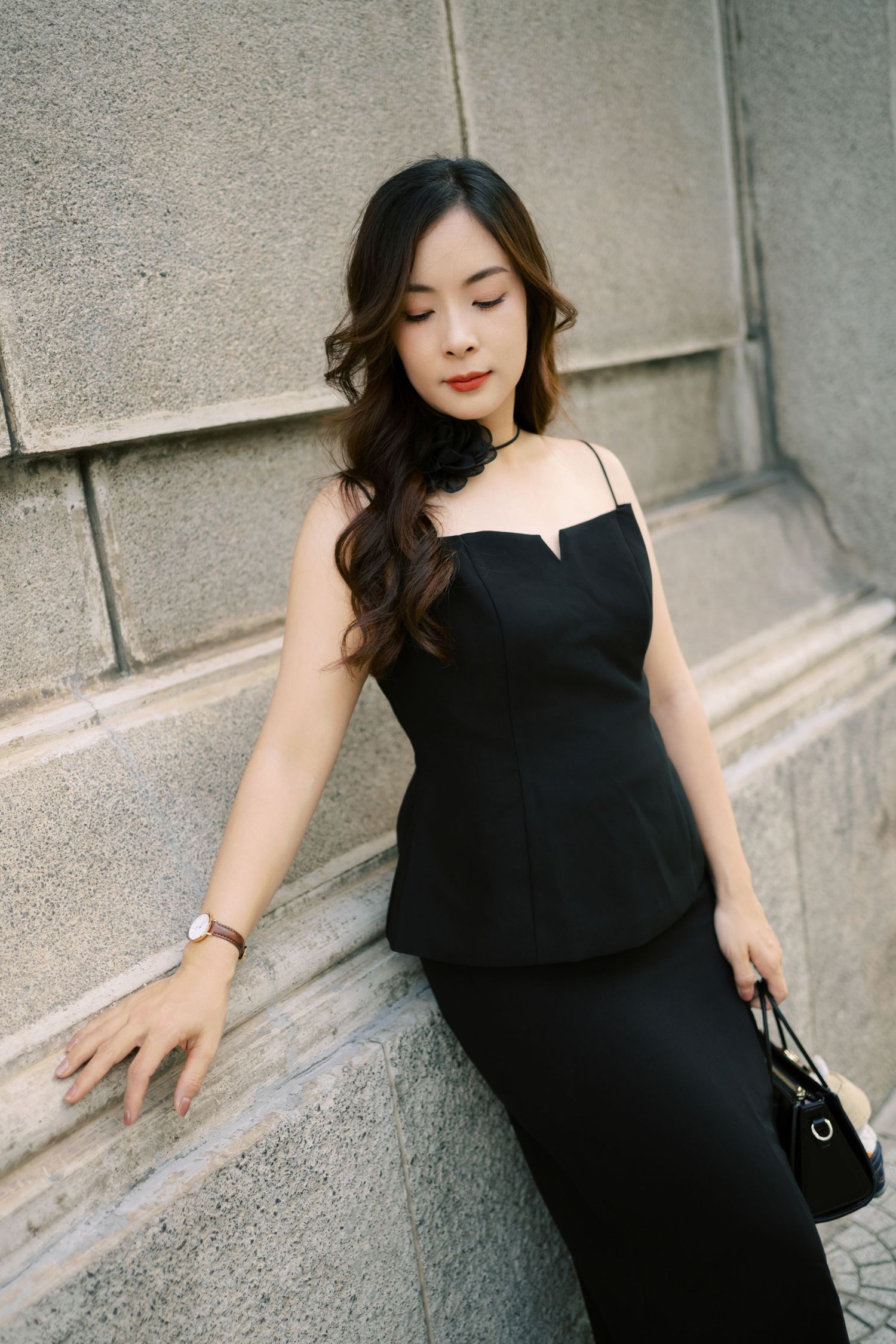 Elegant figure in black evening dress poses gracefully against stone wall while holding designer purse.