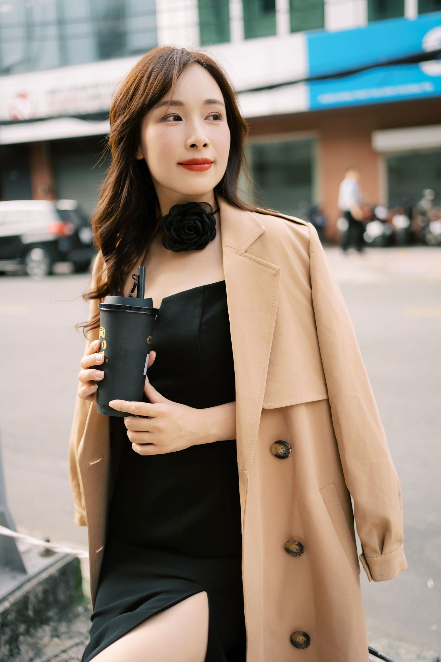 A stylish woman in a camel coat and black dress holds a dark coffee cup while standing on a city street.