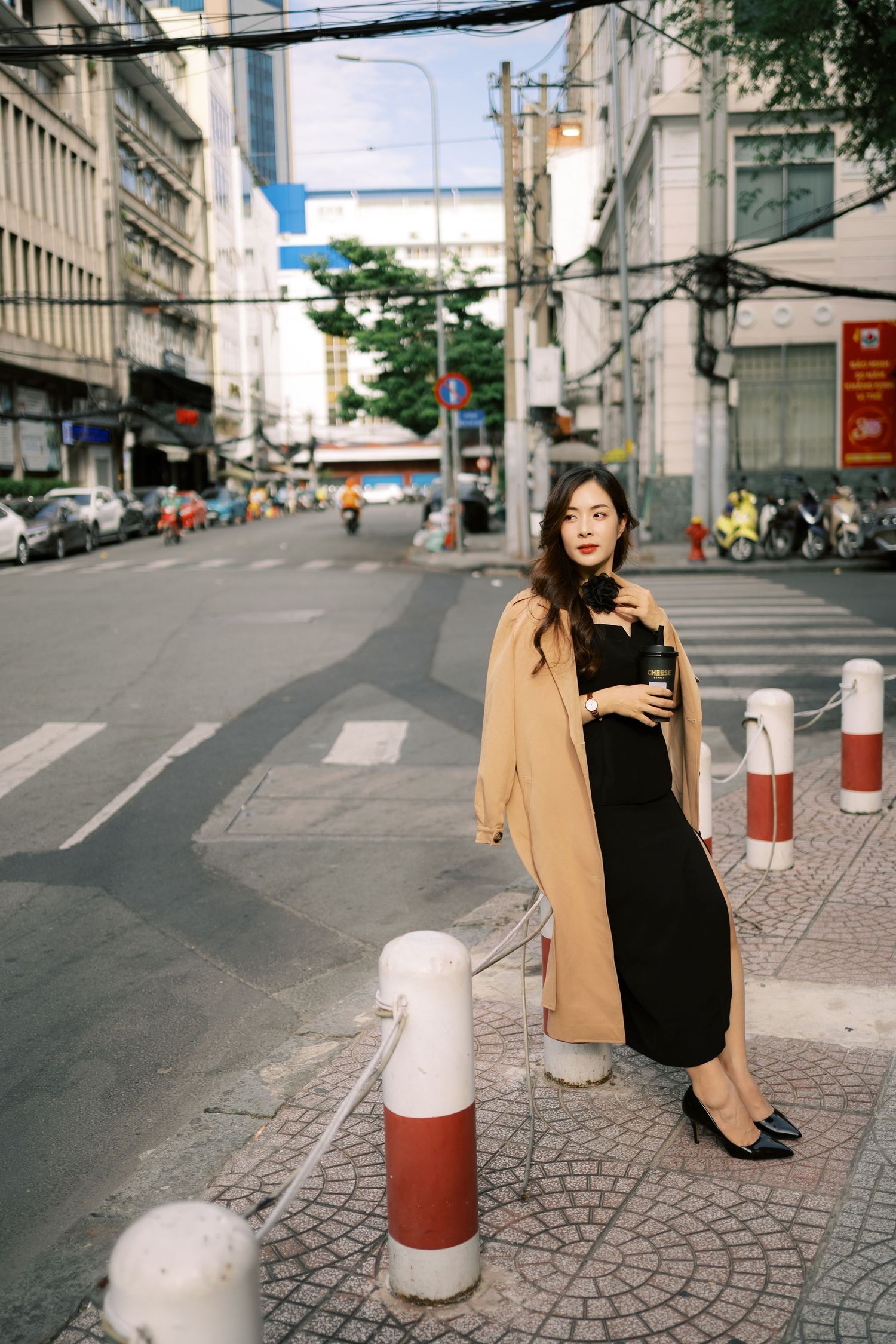 A street scene in an Asian city with red and white bollards on the sidewalk and someone in a beige coat and black dress.