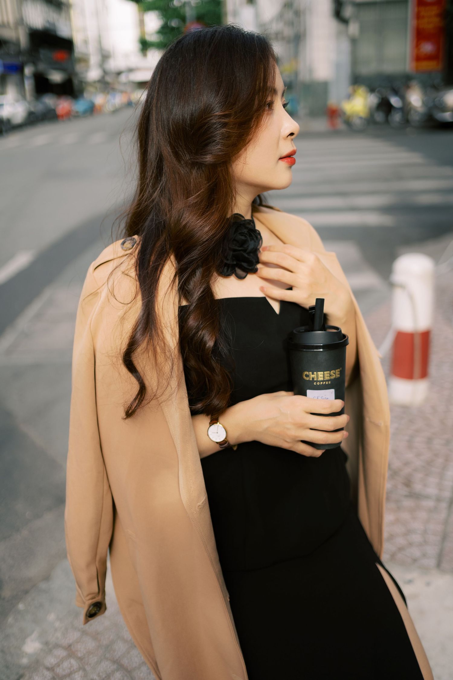 Stylish woman in black dress and beige coat holding coffee cup on city street with elegant side profile pose.