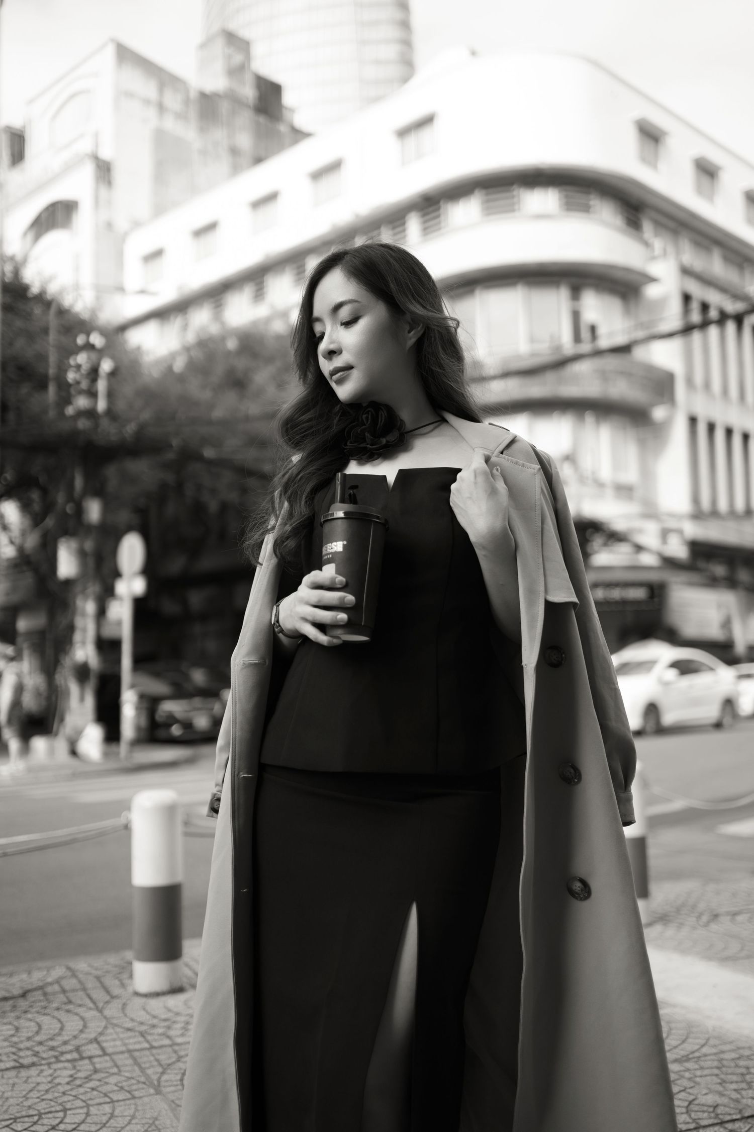 Black and white street photography of a woman in a long coat holding a coffee cup against an urban backdrop.
