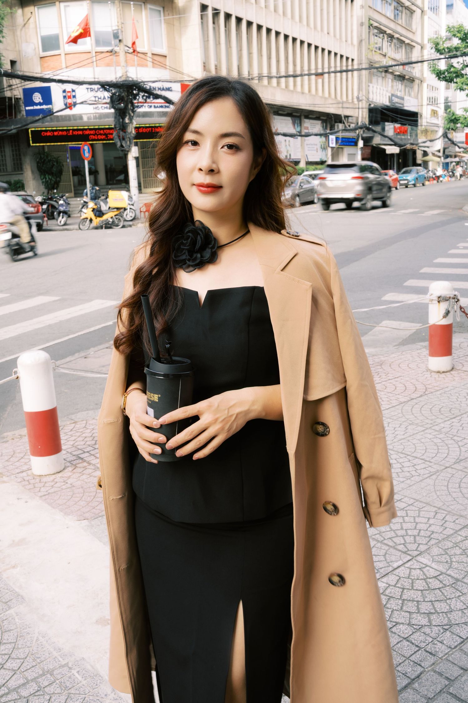 Stylish person in black dress and tan trench coat holding coffee cup on busy city street in Vietnam.