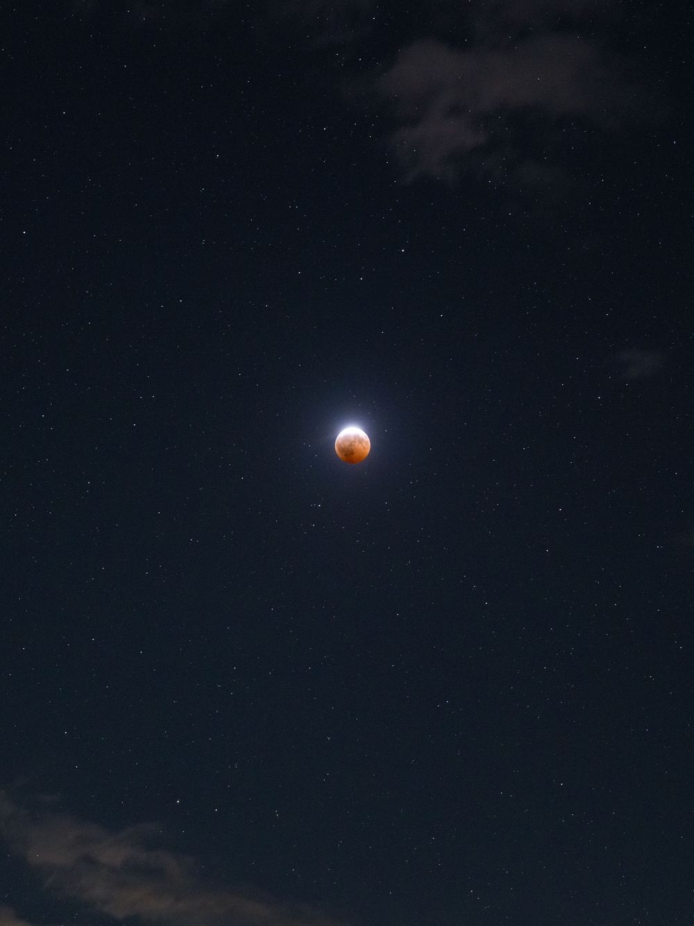 A stunning lunar eclipse with blood moon glowing orange-red against a dark starry night sky with wispy clouds.