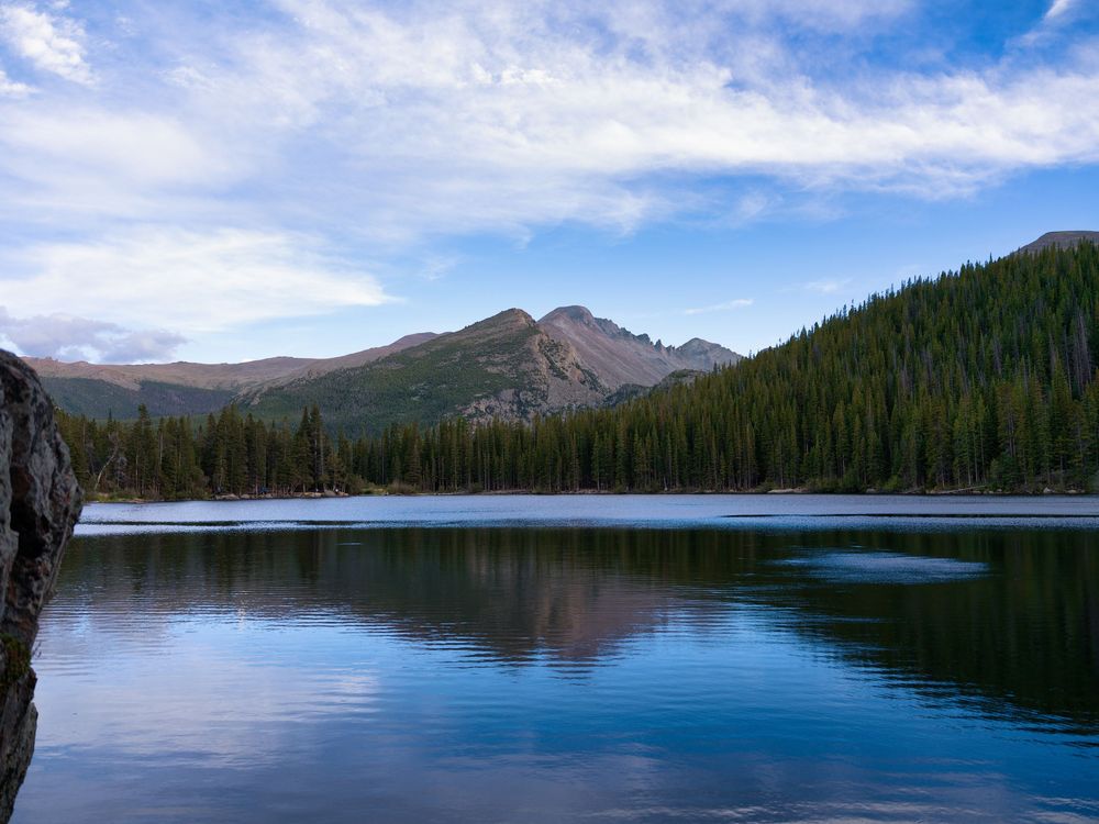 Serene mountain lake reflecting evergreen forests and rocky peaks under a partly cloudy blue sky.