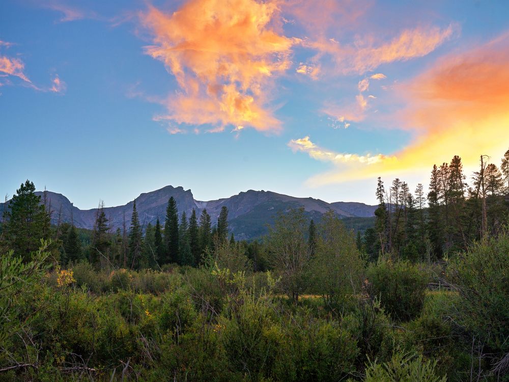 Stunning sunset over Rocky Mountain landscape with colorful clouds, pine trees, and rugged peaks in the background.