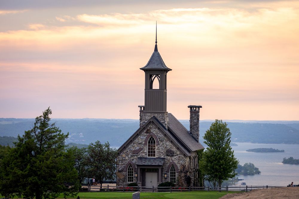 Panoramic view of a small chapel on a hilltop during sunset with mountains and water in the background. Top of the Rock in Branson, Missouri