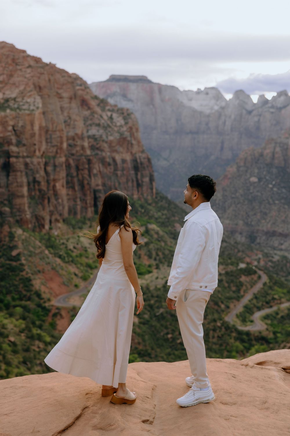 Two people in white outfits stand together on a red rock overlook with dramatic canyon cliffs in Zion National Park.