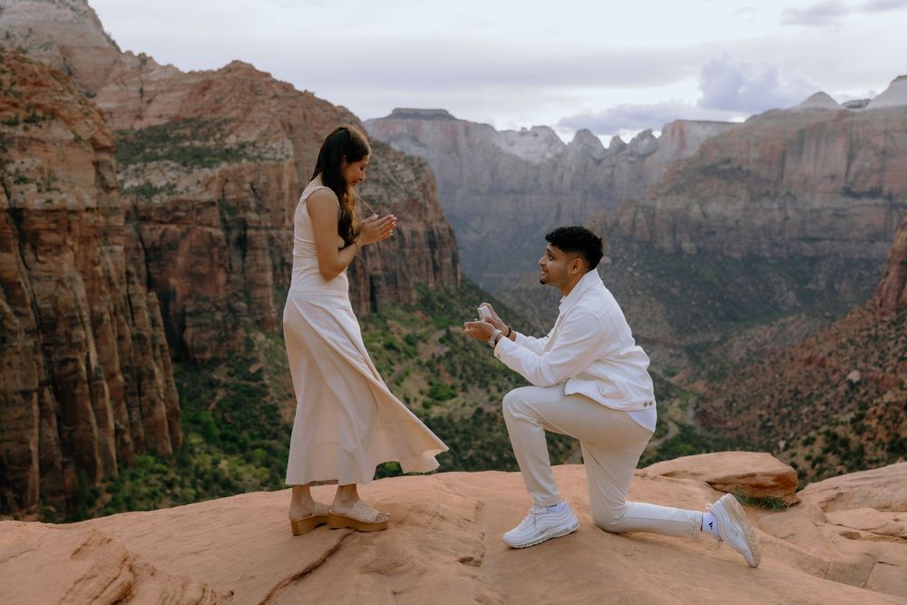 A romantic marriage proposal takes place on red rock cliffs overlooking a dramatic mountain canyon landscape.