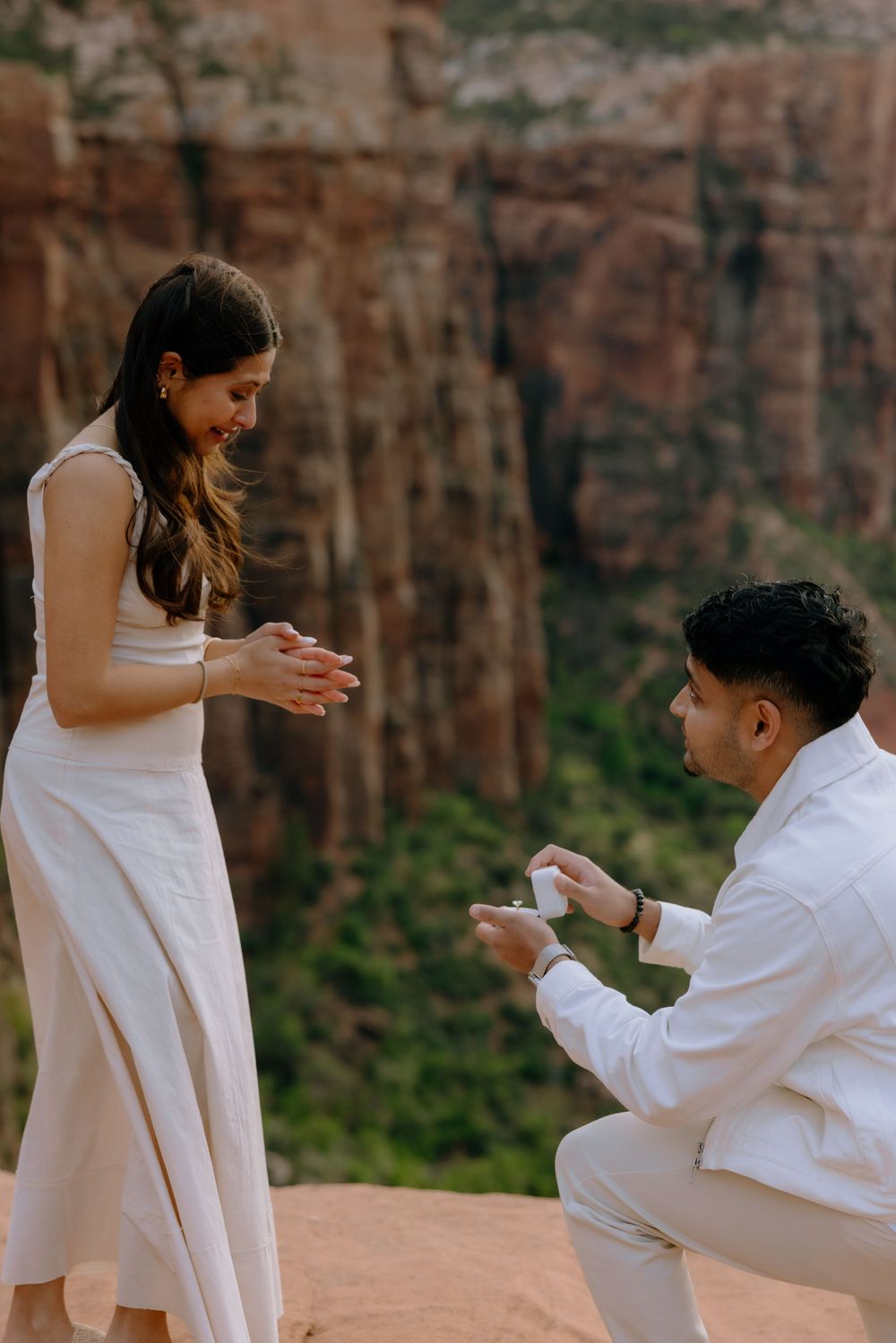A couple shares an emotional moment during their proposal on red rocks with dramatic canyon cliffs in the background.