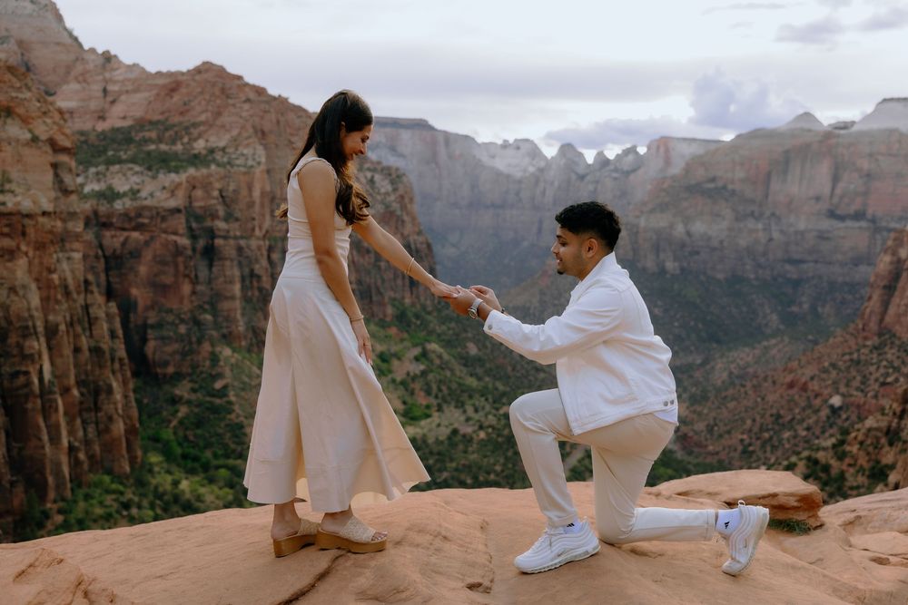 Two people share a romantic proposal moment on a rocky cliff overlooking the majestic canyons of Zion National Park.