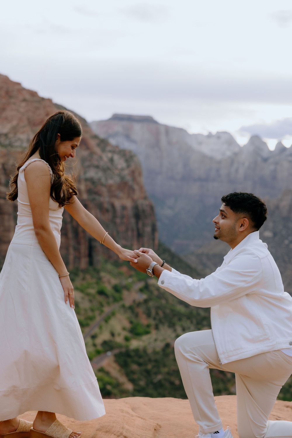 A romantic proposal moment with mountains in Zion National Park as a couple stands on red sandstone rocks at sunset.