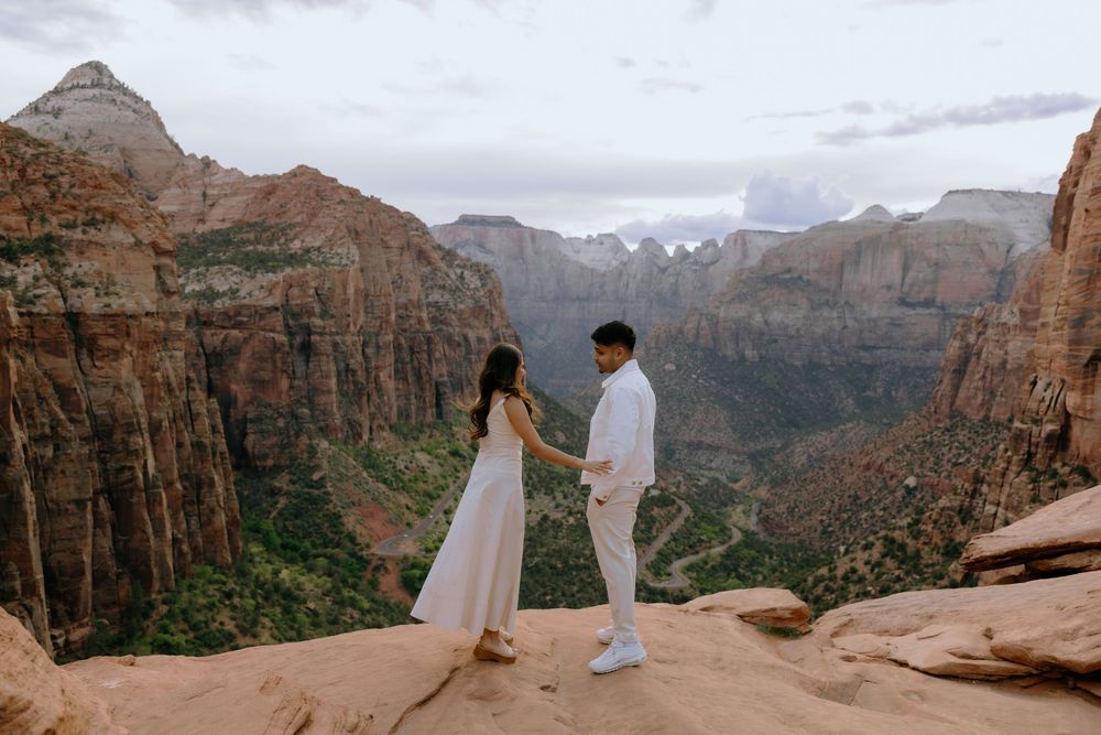 Two people in white attire stand together on red rock overlook against dramatic canyon landscape in Zion National Park.
