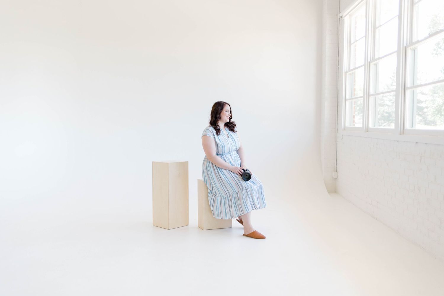 Someone wearing a light blue dress sits on a wooden pedestal in a bright white minimalist room near a window.