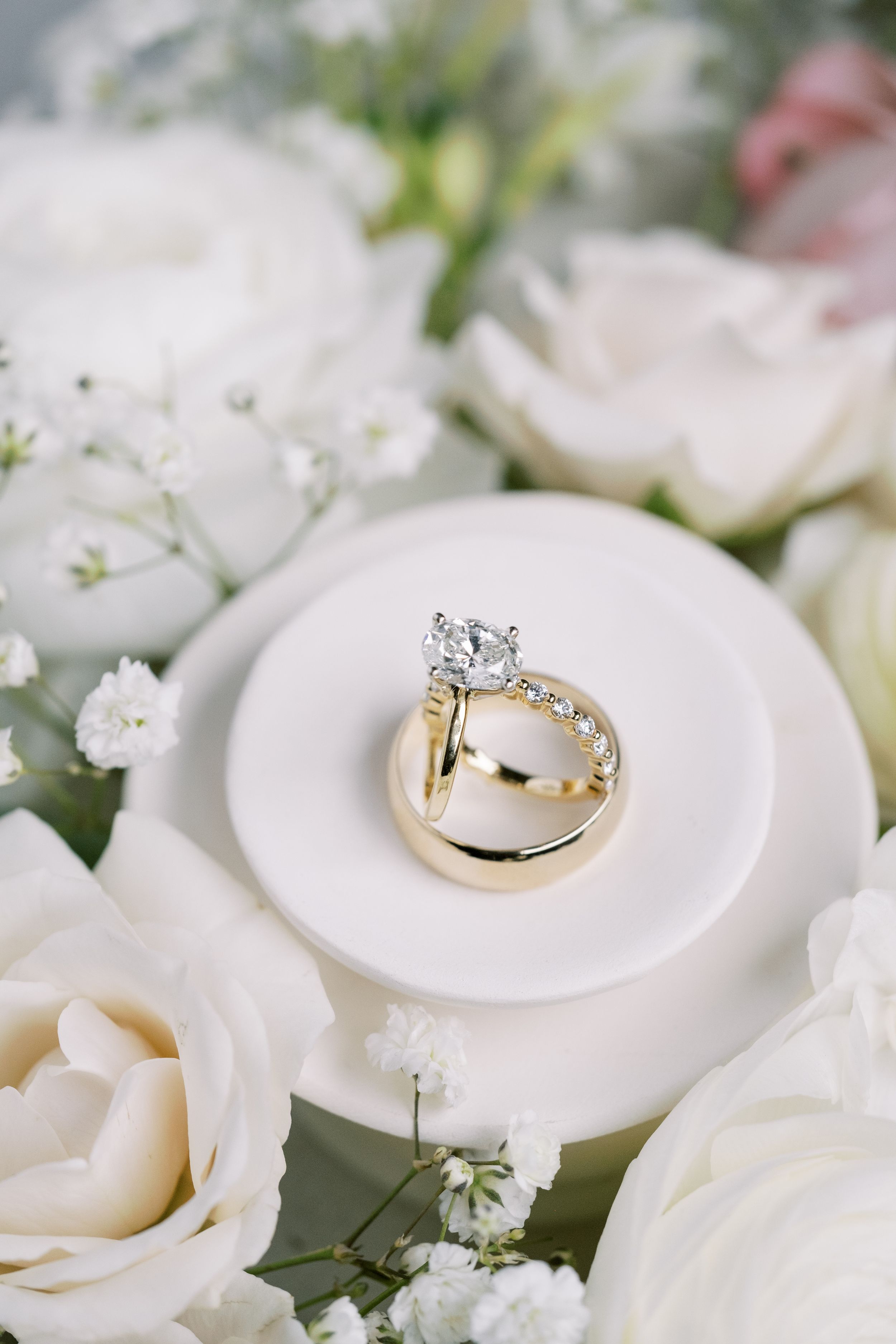 Wedding rings displayed on a white ceramic dish surrounded by delicate white roses and baby's breath flowers.