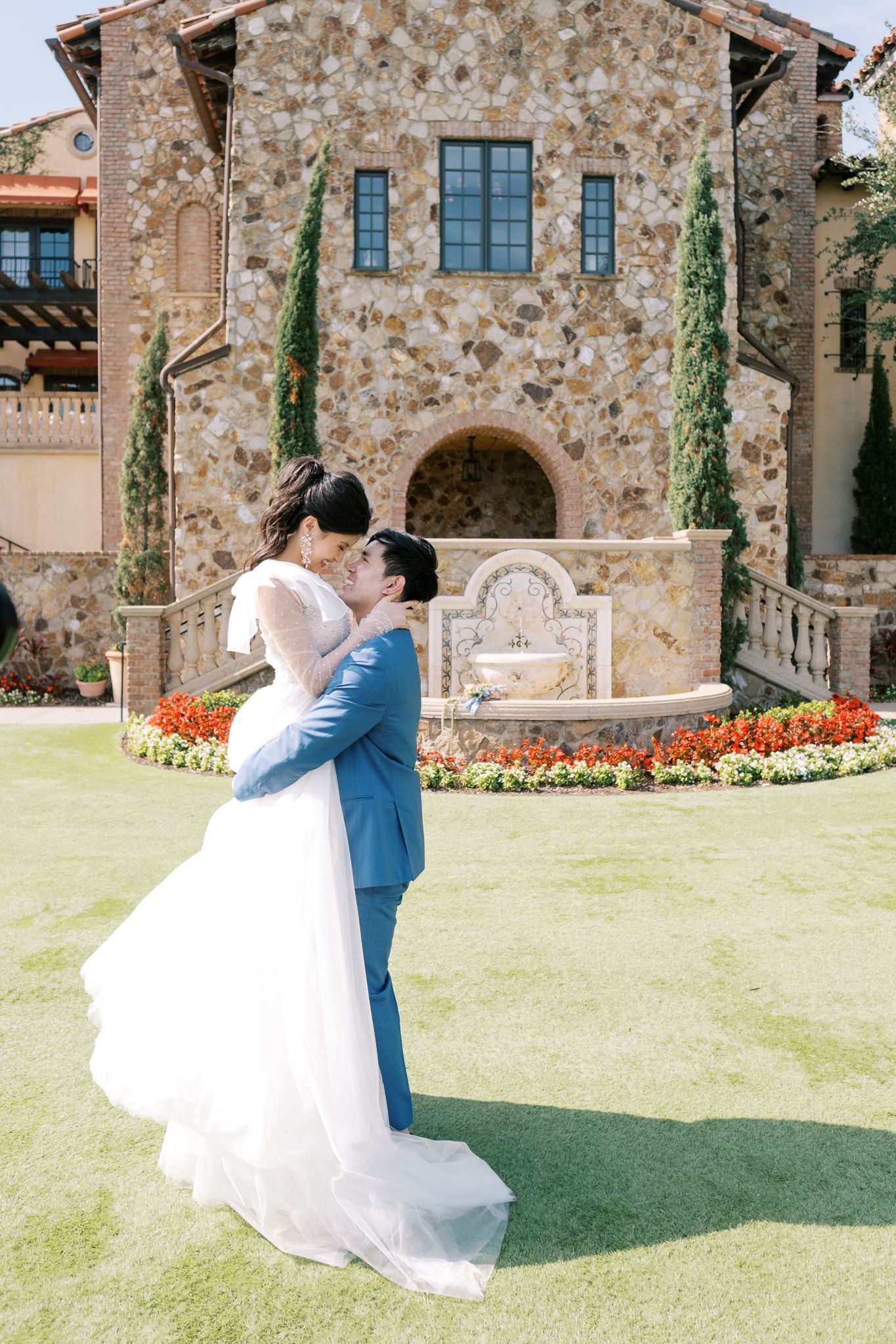 A couple embraces on a manicured lawn in front of a Tuscan-style stone villa with flowering gardens.