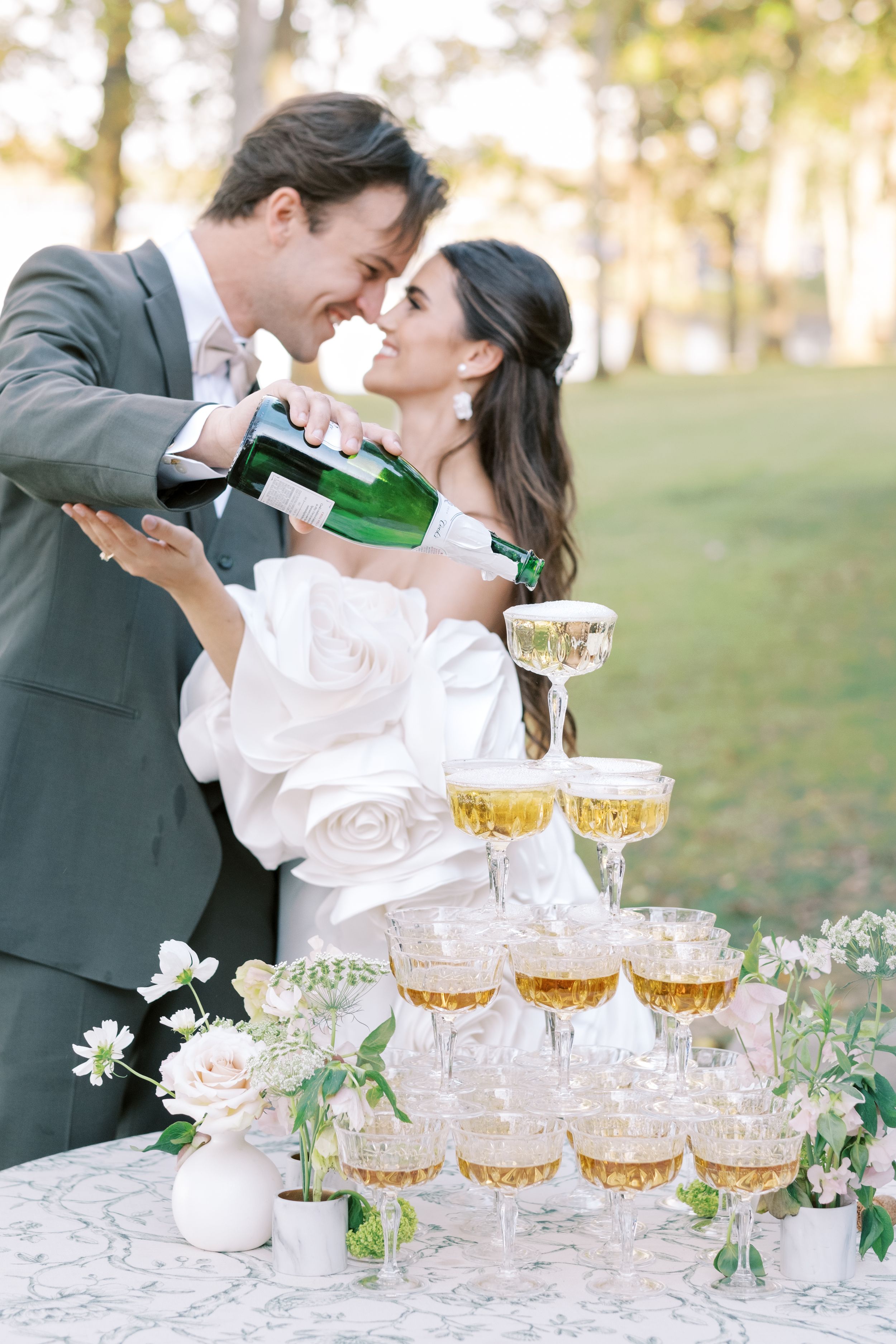 A couple pouring champagne into a tower of coupe glasses decorated with delicate white flowers on an outdoor table.