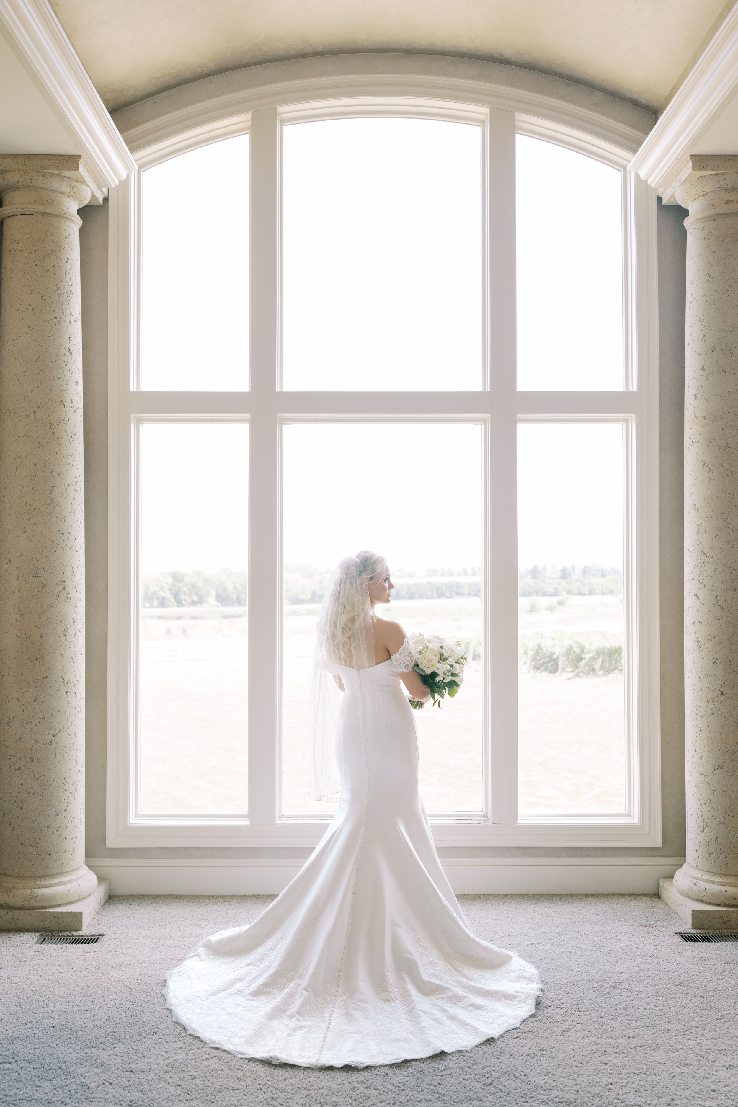 Elegant white wedding dress with long flowing train displayed in grand columned room with arched windows in natural light.
