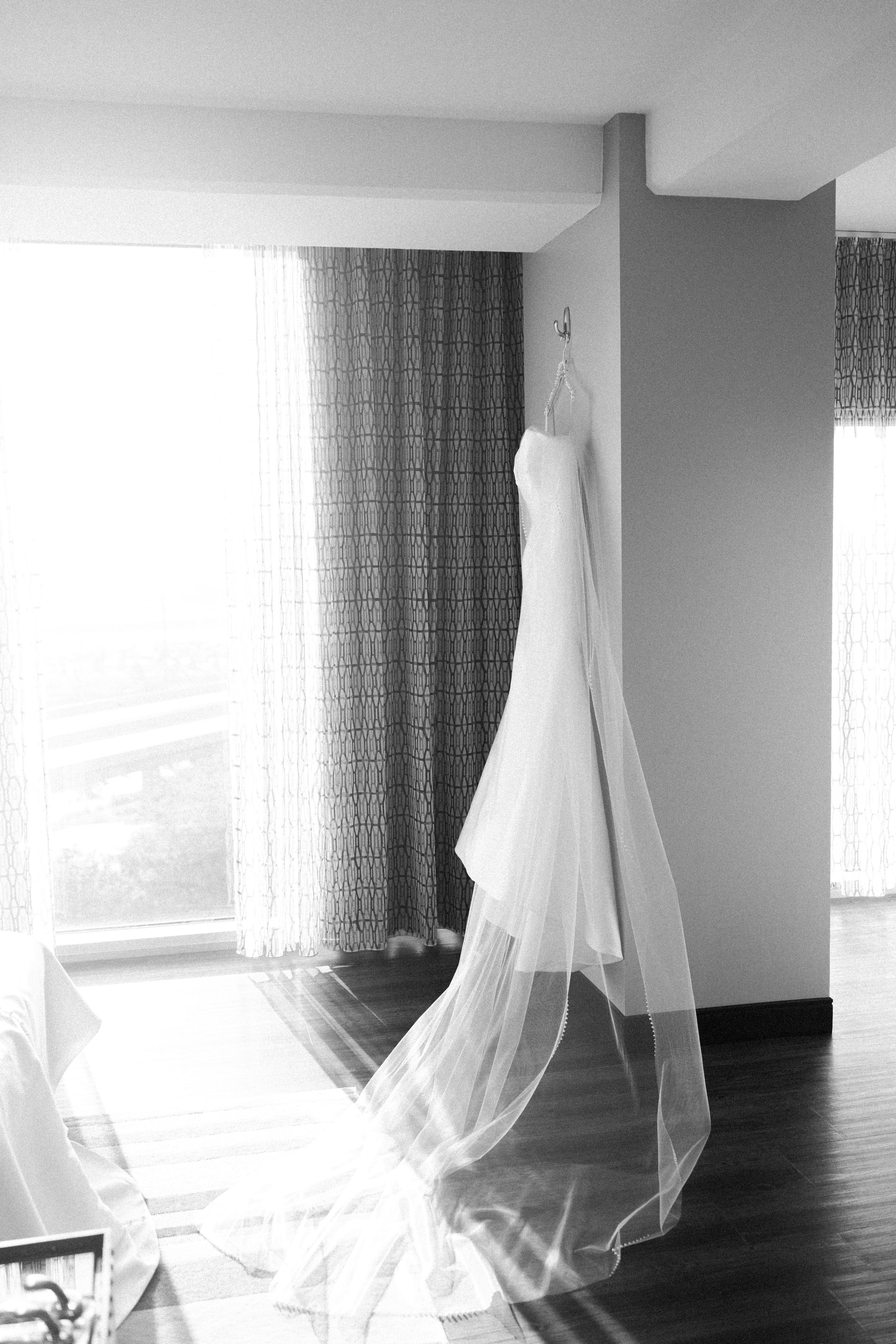 A white wedding dress with flowing veil hangs elegantly against a window with patterned curtains in a sunlit room.