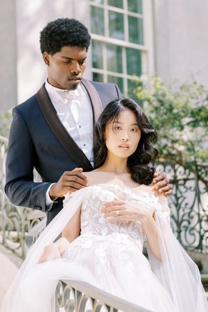 Couple in formal wedding attire pose elegantly on outdoor stairs with ornate railing and greenery in background.