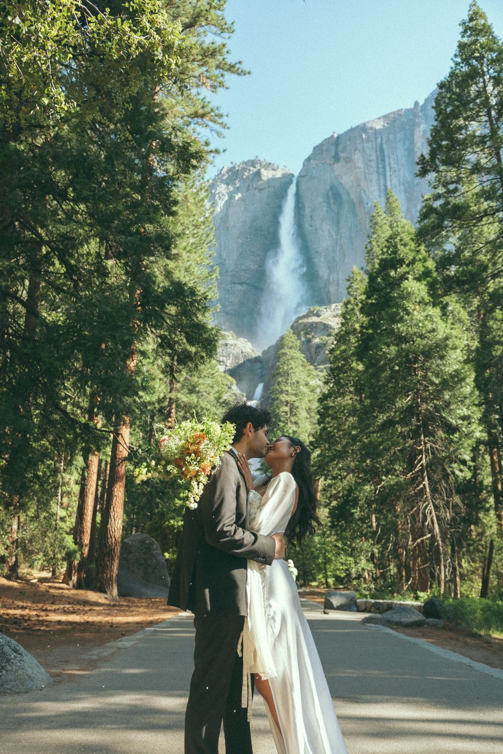 A couple embraces and kisses on a paved path in Yosemite National Park with Yosemite Falls cascading in the background.