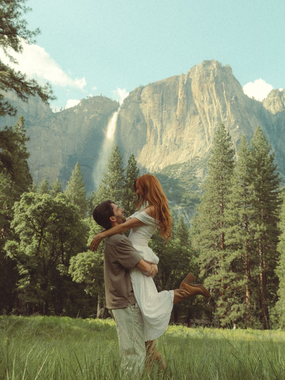 Two people dance and embrace together in a meadow with towering mountain cliffs and evergreen trees in the background.