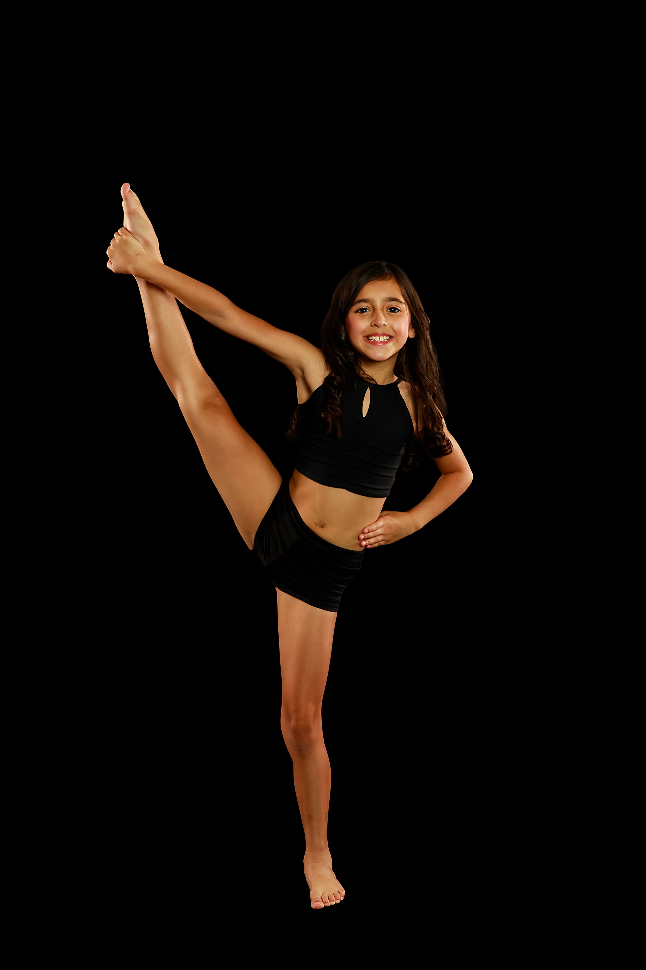 Athletic dancer performs high kicks in black outfit against dark background.