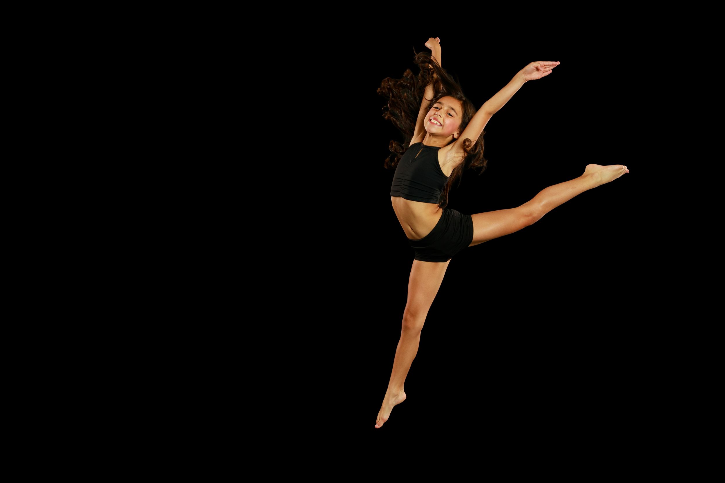 Dancer in black performing a graceful leap against dark background showing impressive flexibility and form.