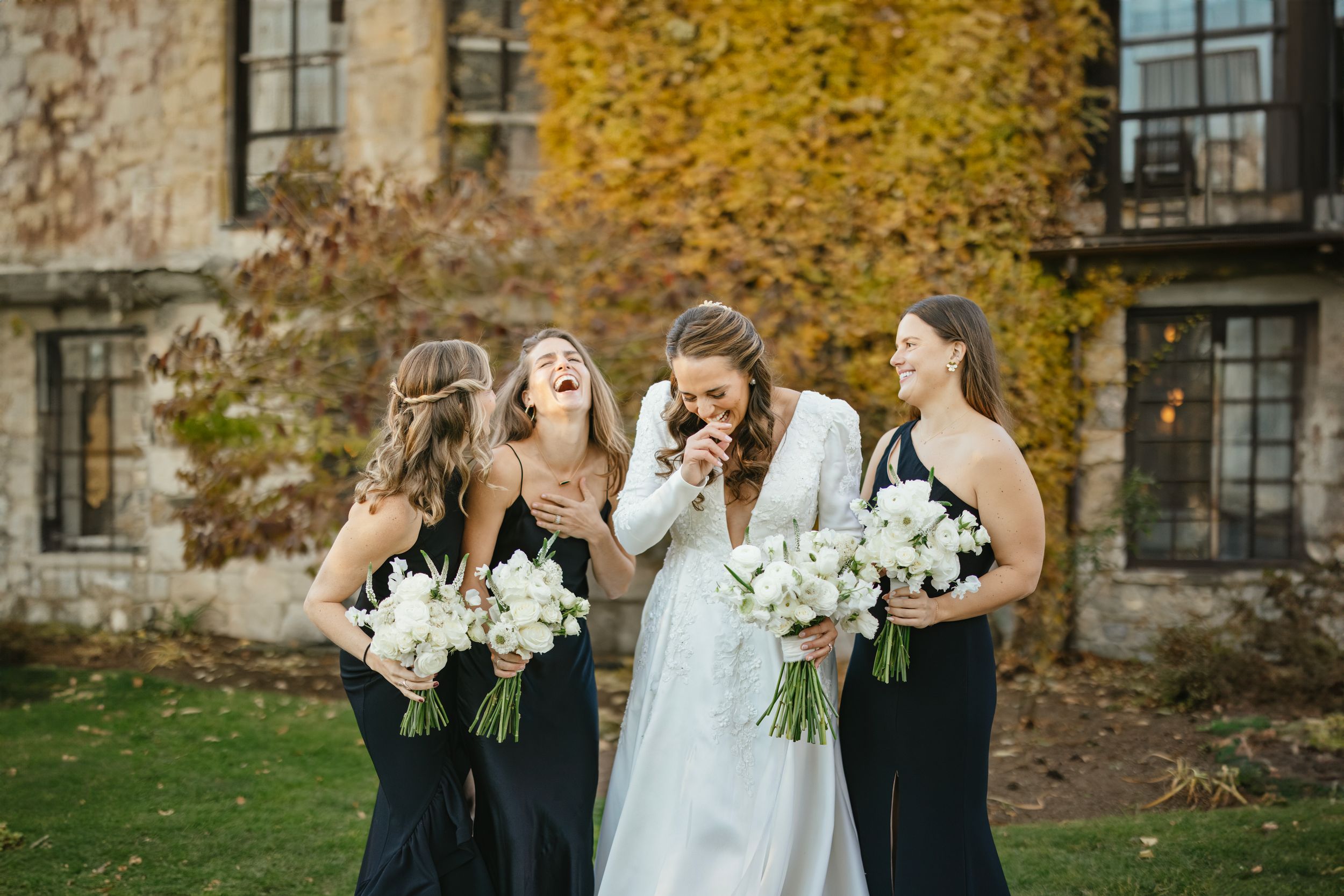 Bride and bridesmaids in navy dresses share laughter while holding white floral bouquets against autumn stone building.