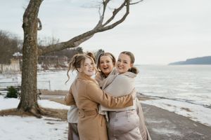Three friends share joyful embrace while standing on snowy beach shoreline during winter day.