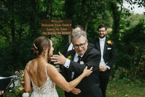 Emotional embrace between a bride in a lace dress and guest at an outdoor wedding ceremony with wooden welcome sign.