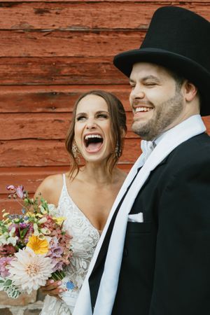 Newly married couple sharing a joyful laugh against rustic red barn wall with colorful wildflower bouquet.