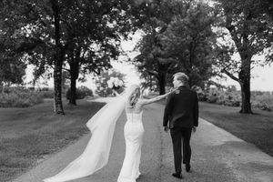 Black and white photo of a couple walking down a tree-lined path, with a flowing wedding veil catching the breeze.